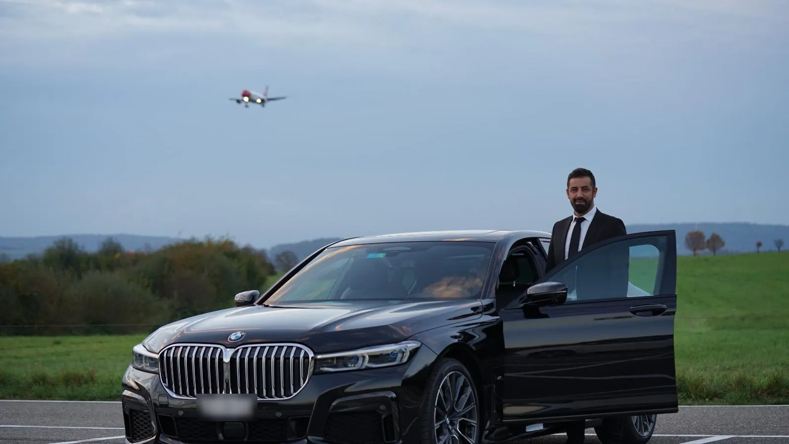Chauffeur in a dark suit at the open door of a black sedan, a plane landing in the background